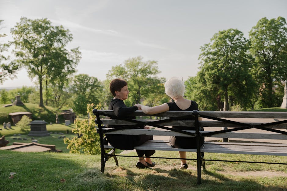 Two senior women sitting on a bench in a serene cemetery setting, offering support.